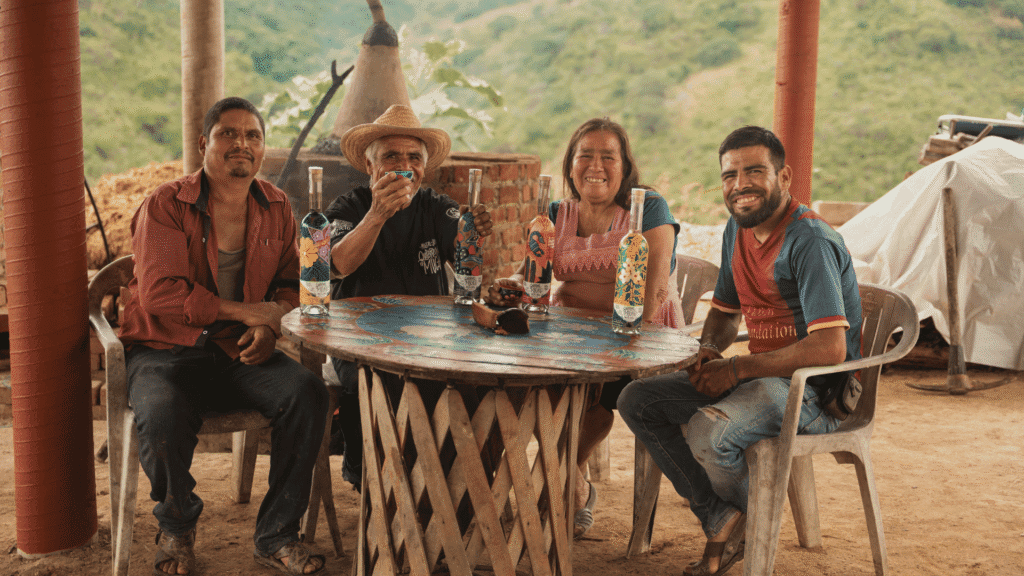 mezcal artesanal de Oaxaca sobre mesa de madera, agaves al fondo